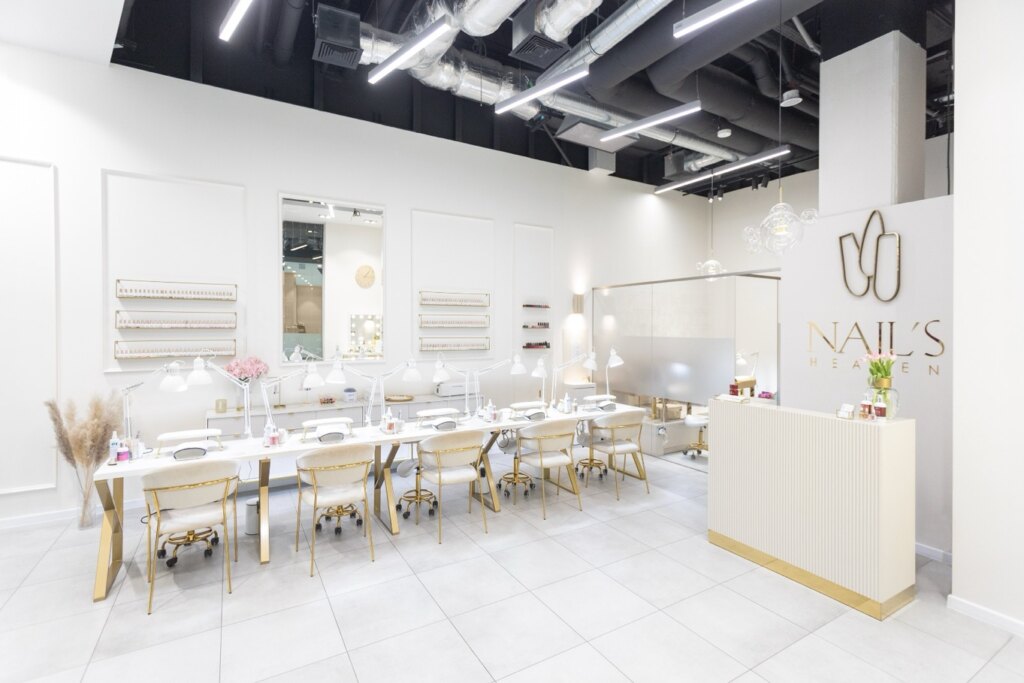 Salon paznokci Nails Heaven w Katowicach - pięć stanowisk do manicure z białymi fotelami i złotymi detalami, ściana z lakierami hybrydowymi i recepcja z logo salonu.